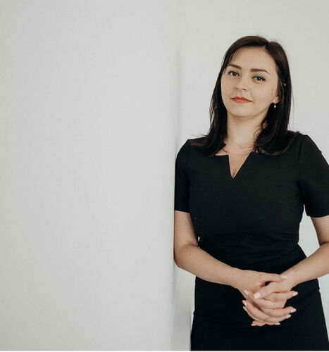 Femeie în rochie neagră sprijinită de un perete alb, cu o expresie calmă și încrezătoare.