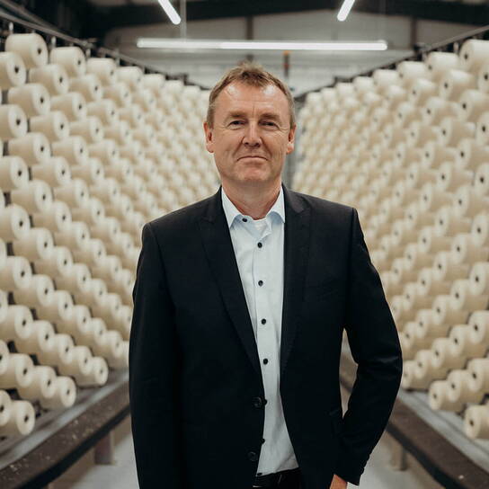 A man in a suit stands confidently in a textile factory surrounded by rows of large spools.