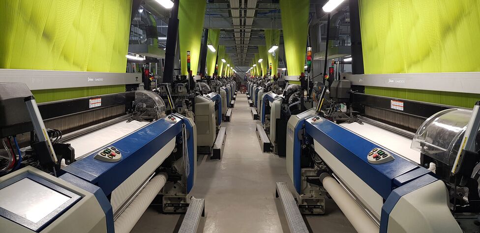 Mașini industriale de țesut aliniate într-o fabrică modernă, sub lumina reflectată de perdelele verzi.