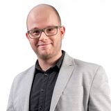 Ein Mann in einem grauen Blazer und schwarzem Hemd lächelt selbstbewusst in die Kamera.