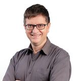 Ein Mann mit Brille und grauem Hemd lächelt selbstbewusst mit verschränkten Armen vor einem weißen Hintergrund.