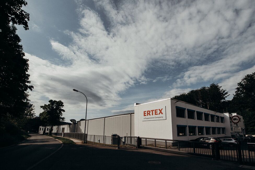 A modern industrial building labeled "ERTEX" with a clear sky and trees in the background.