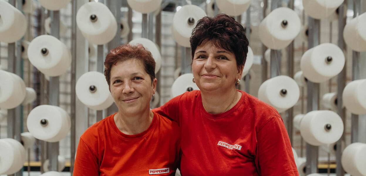 Două femei zâmbitoare în tricouri roșii stau în fața unor role mari de ață într-o fabrică modernă.