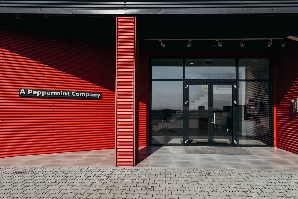 Intrarea modernă a unei clădiri cu fațadă roșie și semn "A Peppermint Company" este luminată de soare.