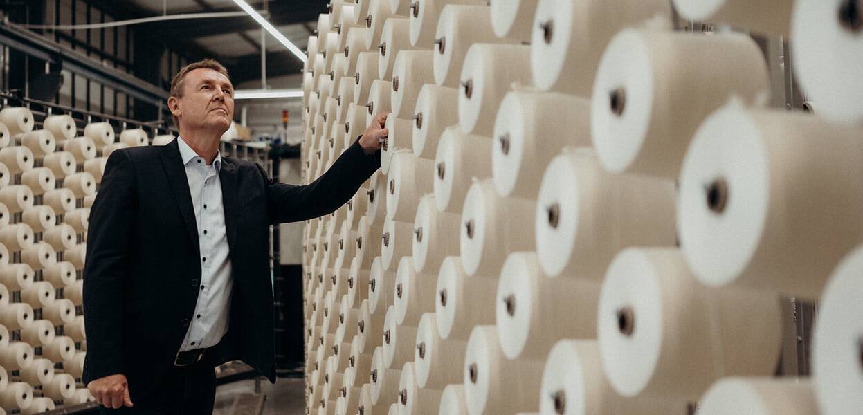 A man in a suit examines rows of large white spools in a textile manufacturing facility.