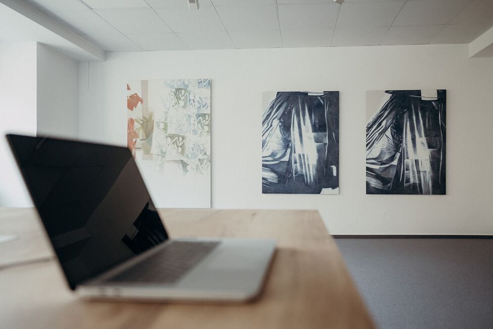 A laptop sits on a wooden table in a minimalist gallery displaying abstract artwork on the walls.