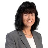 Eine Frau mit dunklem Haar und grauem Blazer lächelt vor einem weißen Hintergrund in die Kamera.