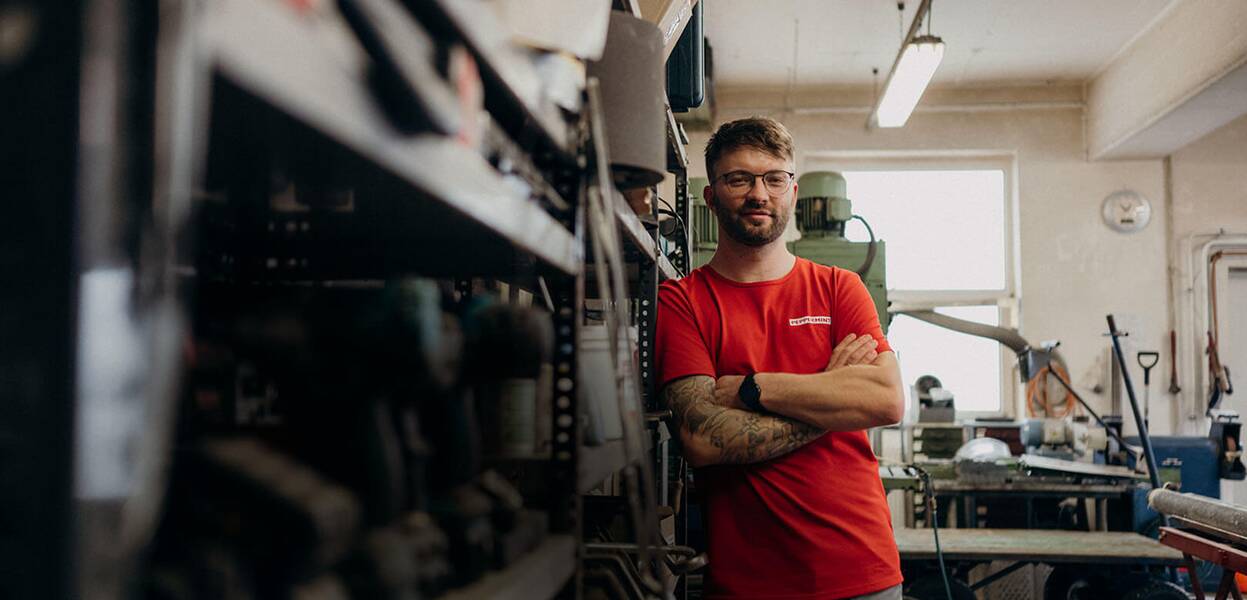 A man in a red shirt stands confidently in a workshop, surrounded by tools and machinery, showcasing his expertise.