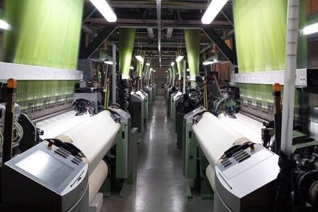 A row of industrial weaving machines operates in a textile factory, producing fabric under bright fluorescent lights.