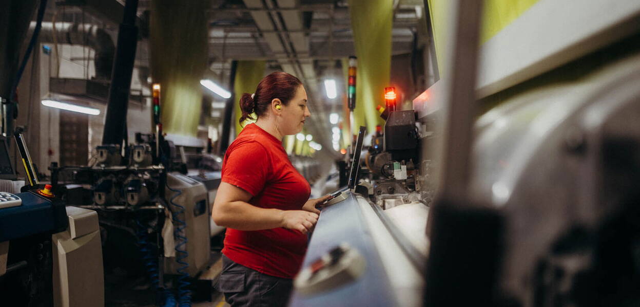 O femeie lucrează concentrată la o mașinărie industrială într-o fabrică, purtând un tricou roșu.