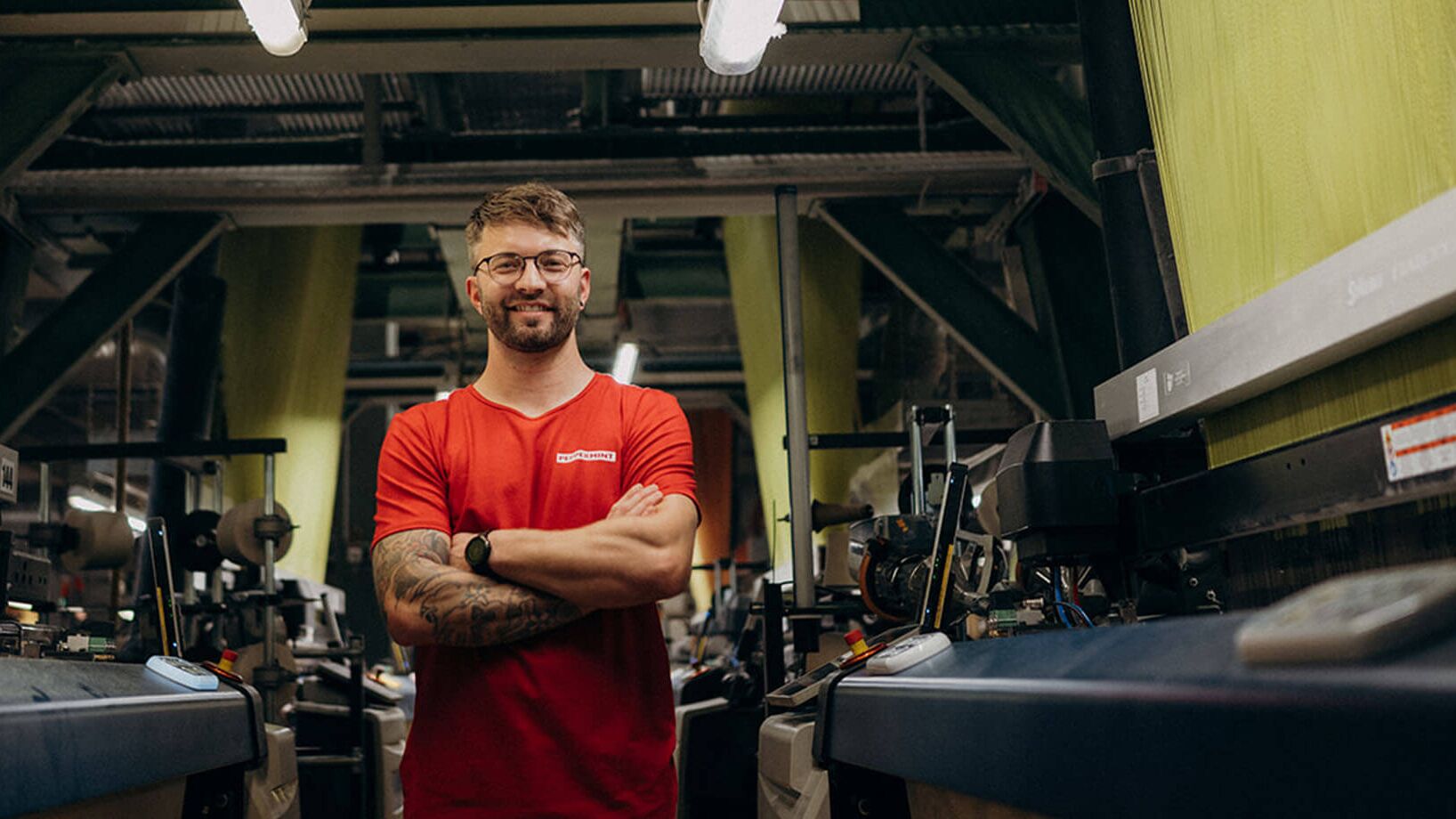 Ein Mann in rotem Shirt steht selbstbewusst in einer modernen Fabrikhalle zwischen Maschinen.