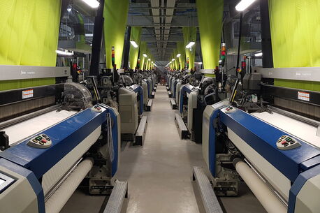 Rows of advanced textile machines in a factory are busy weaving fabric under bright lights.