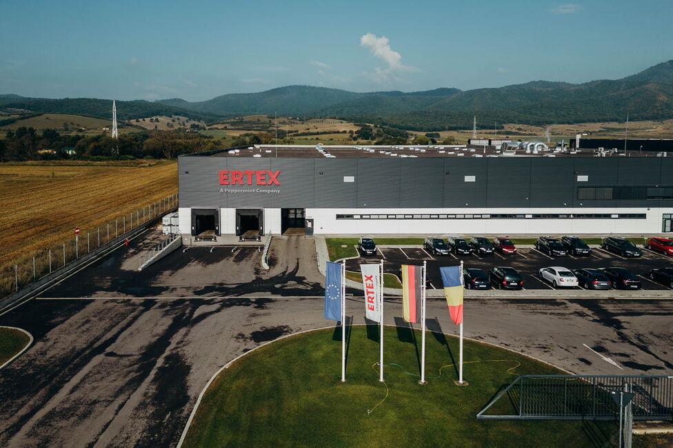Clădirea Ertex International se află într-un peisaj rural, cu mașini parcate și steaguri în față.