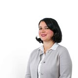 Femeie zâmbitoare în sacou gri stă în picioare, emanând profesionalism și încredere pe un fundal alb.