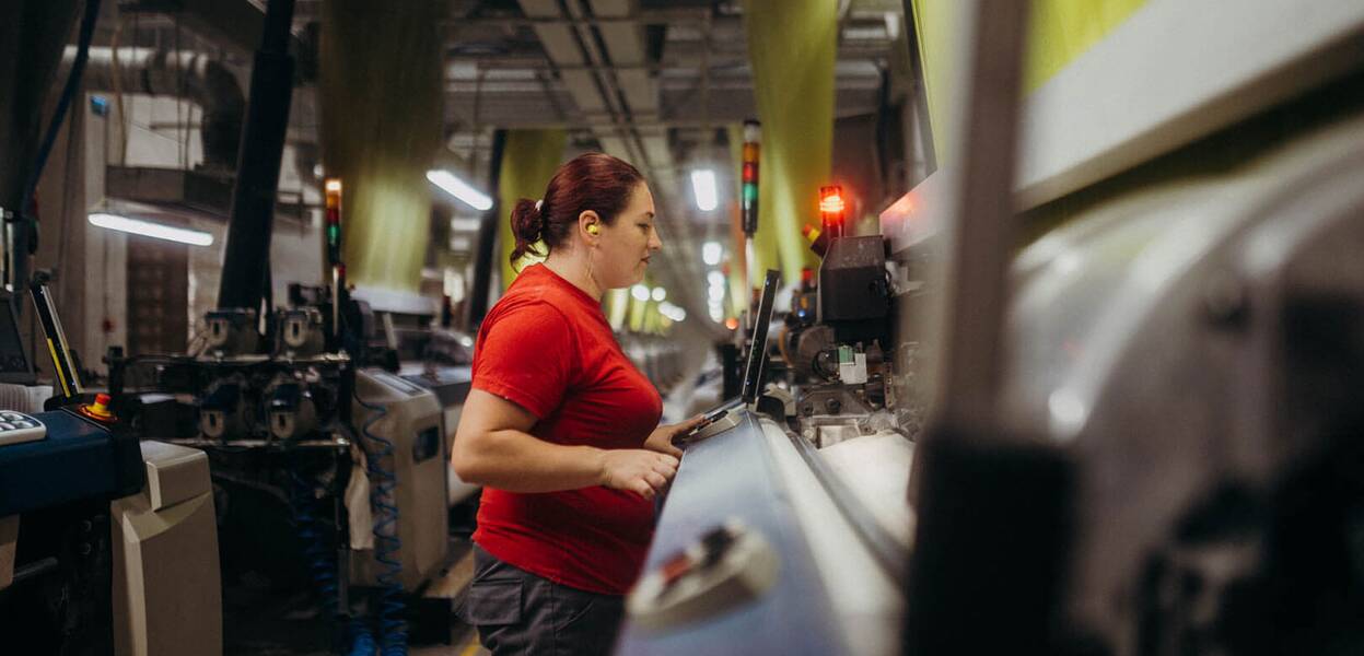 O femeie lucrează concentrată la o mașinărie industrială într-o fabrică, purtând un tricou roșu.