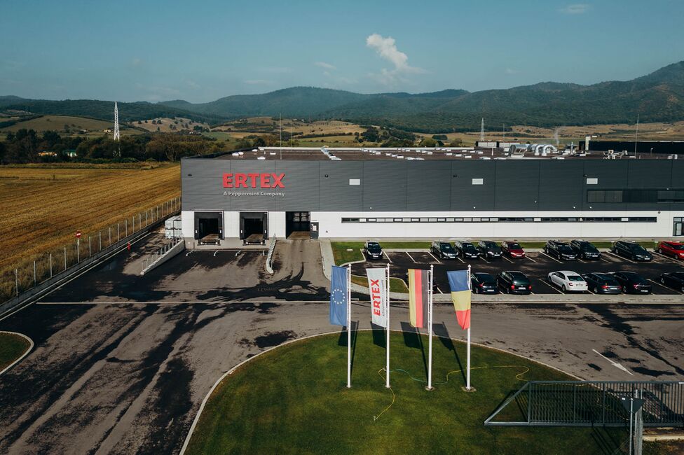 Clădirea Ertex International se află într-un peisaj rural, cu mașini parcate și steaguri în față.