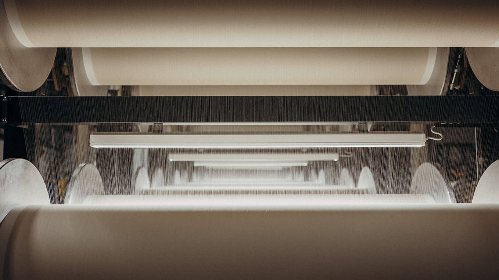 Large industrial textile rollers are aligned in a factory, with fabric being processed under bright lights.