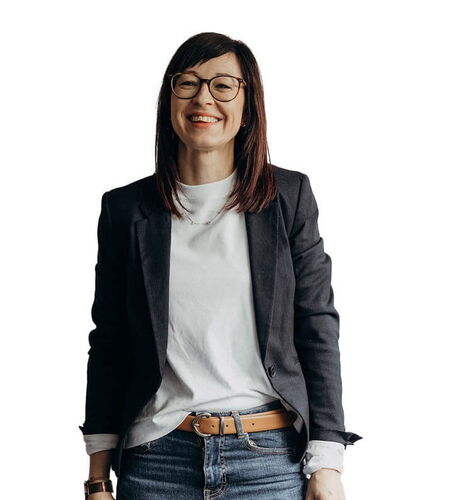 Eine Frau in Jeans und Blazer lächelt freundlich vor einem weißen Hintergrund.