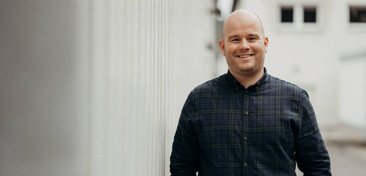 A smiling man in a plaid shirt stands outdoors against a blurred background, exuding confidence and positivity.