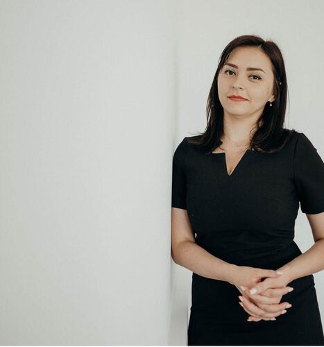 Femeie în rochie neagră sprijinită de un perete alb, cu o expresie calmă și încrezătoare.