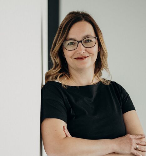 A woman in glasses and a black dress stands confidently with arms crossed, leaning against a white wall.