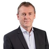 Ein Mann in einem eleganten Anzug steht vor einem weißen Hintergrund und blickt direkt in die Kamera.